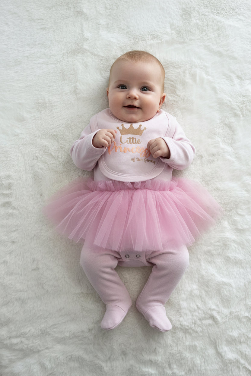 A baby girl's pink footed sleeper set with a matching bib, laid flat on a white textured blanket. The sleeper is light pink with a darker pink tutu skirt and features gold foil text reading 'Little Princess' with a crown.
