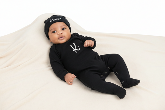 Baby wearing a black outfit with a logo and a matching hat on a white blanket