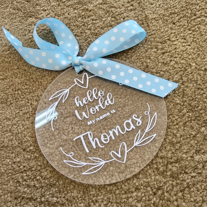 A personalized clear acrylic round ornament with white text reading 'hello World MY name is Thomas' and white leaf heart designs, tied with a light blue polka dot ribbon on a beige carpet background.