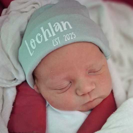 Newborn baby wearing a hat with 'Lochlan EST 2025' text, wrapped in red blanket.