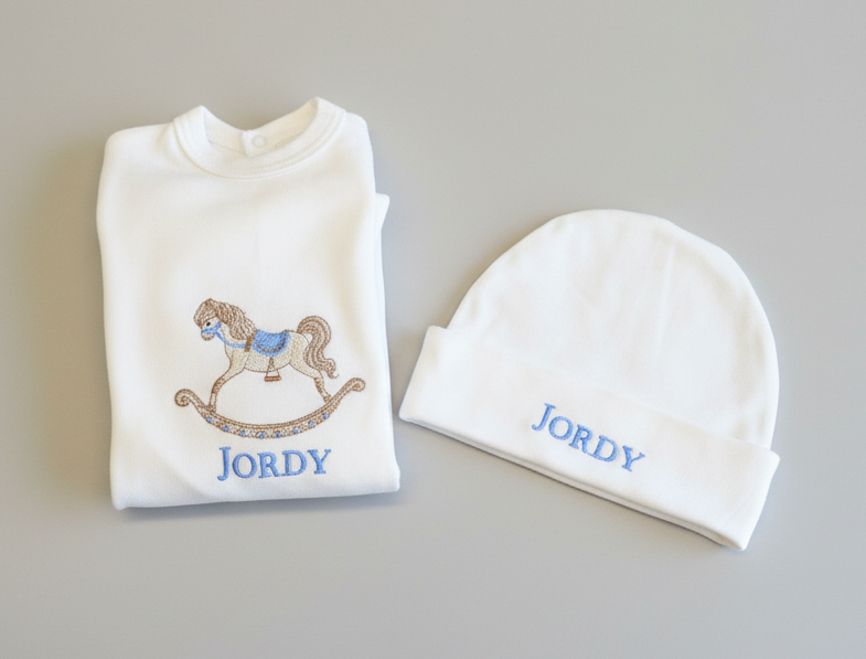 Folded baby outfit with embroidered rocking horse and 'Jordy' text on a textured surface